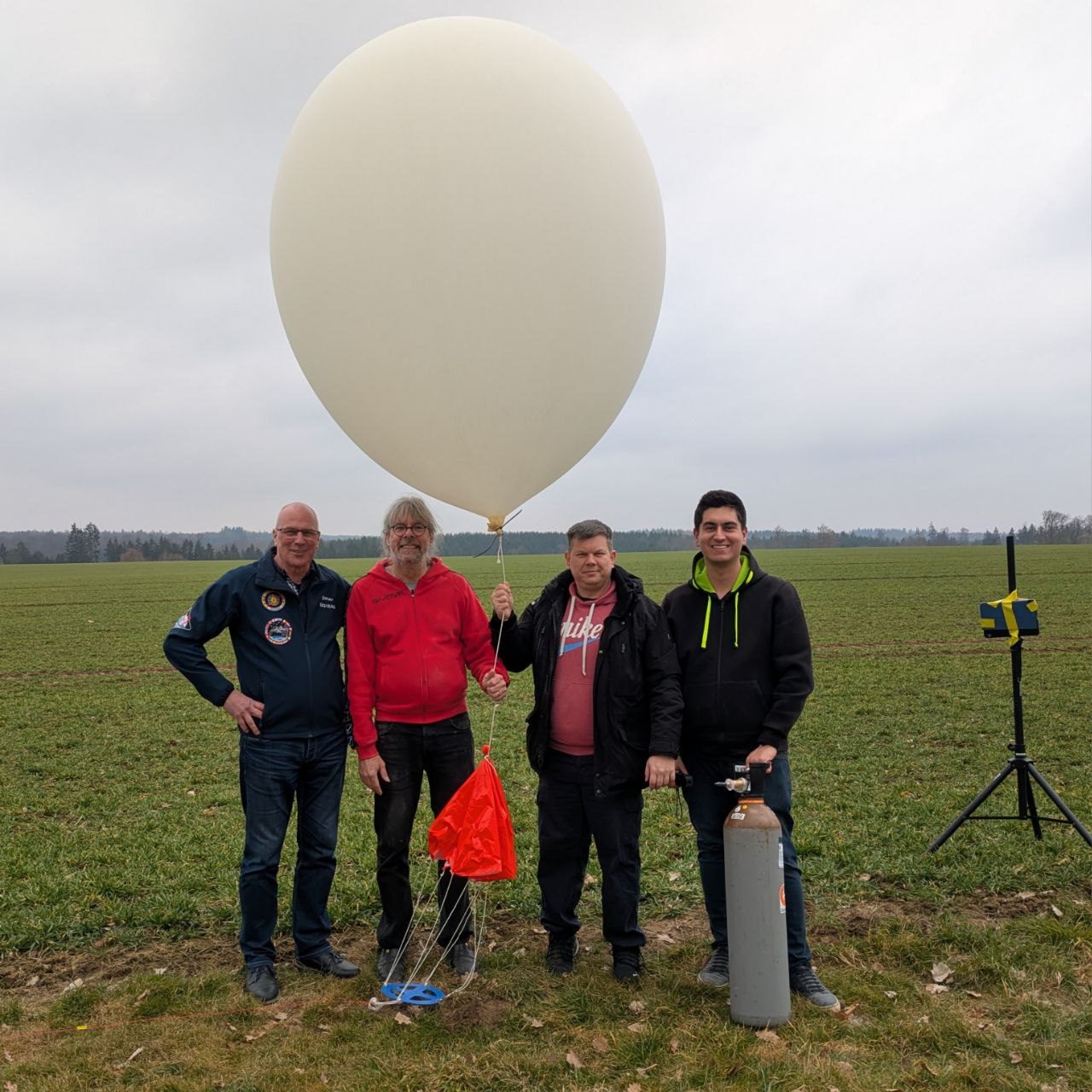 Vier Funkamateure stehen auf einem Feld vor dem mit Helium gefüllten Stratosphärenballon. Einer hält die Schnur, daneben liegen Fallschirm und Nutzlast, rechts steht die Heliumflasche.